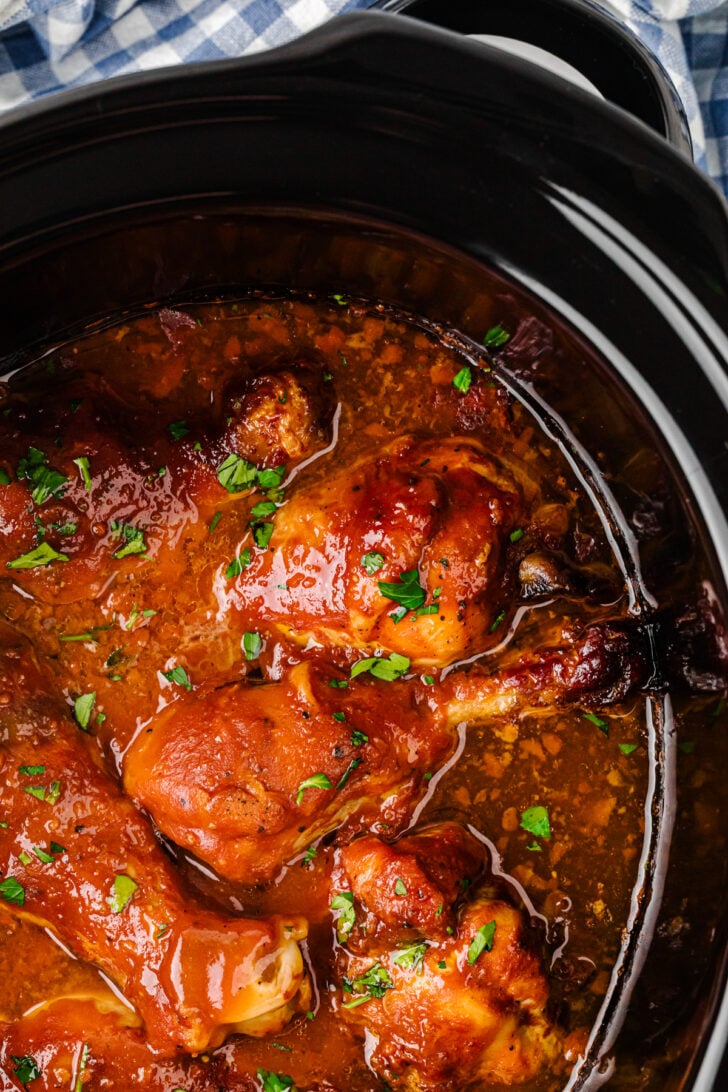 एक धीमी कुकर के अंदर एक अमीर, लाल-भूरे रंग की चटनी में उबालते हुए रसदार चिकन पैरों का क्लोज-अप, कटा हुआ ताजा अजमोद से गार्निश किया गया।