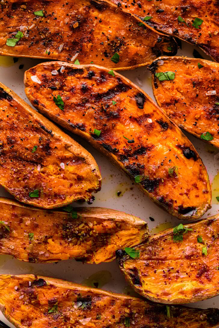 चार्ट ग्रिल के निशान के साथ ग्रील्ड शकरकंद के हिस्सों के क्लोज़-अप, परतदार समुद्री नमक, फटा काली मिर्च और ताजा अजमोद के साथ छिड़के। आलू में कुरकुरी किनारों के साथ एक कारमेलाइज्ड, सुनहरा-नारंगी सतह होती है।