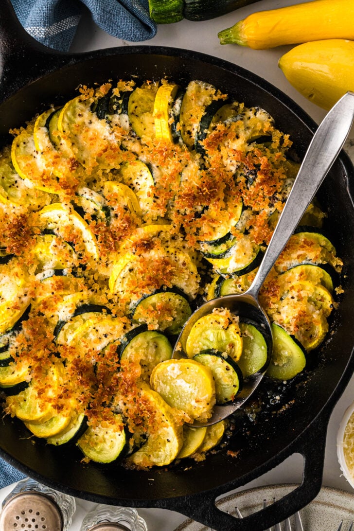 एक पके हुए तोरी और पीले स्क्वैश पुलाव से भरे एक कच्चा लोहे की कड़ाही का ओवरहेड दृश्य, सुनहरा, कुरकुरी ब्रेडक्रंब और पनीर के साथ सबसे ऊपर है। एक सेवारत चम्मच एक हिस्से को स्कूप करता है, जो निविदा, स्तरित सब्जियों का खुलासा करता है।