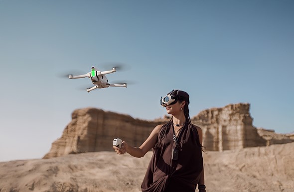 a_woman_stands_in_a_desert_with_goggles_on_her_face_and_a_drone_flying_in_front.jpeg