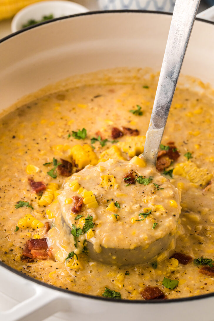 एक सफेद डच ओवन में मलाईदार मकई के चाउडर का क्लोज-अप; एक लाडल स्वीट कॉर्न गुठली, बेकन बिट्स, अजमोद और काली मिर्च के साथ हार्दिक स्कूप को उठाता है।