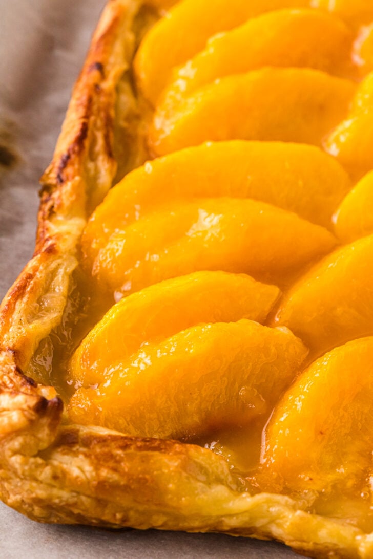 गोल्डन-ब्राउन पफ पेस्ट्री पीच टार्ट ग्लिस्टिंग पीच स्लाइस के साथ पंक्तियों में व्यवस्थित, परतदार क्रस्ट किनारों और चमकदार फल टॉपिंग को दिखाते हैं। एक देहाती प्रस्तुति के लिए तीखा चर्मपत्र कागज पर टिकी हुई है।