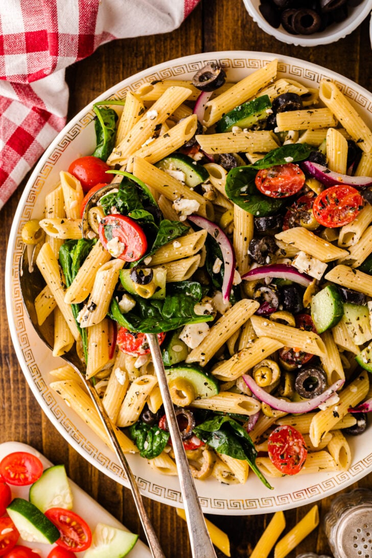 पेनी, चेरी टमाटर, खीरे, काले जैतून, लाल प्याज, पालक, और एक सफेद प्लेट पर फेटा पनीर के साथ भूमध्यसागरीय पास्ता सलाद का ओवरहेड दृश्य, अतिरिक्त सामग्री और एक लाल चेकर नैपकिन से घिरा हुआ है।
