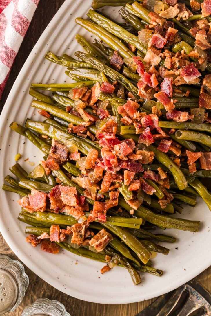 धीमी गति से पकी हुई मेपल बेकन हरी फलियाँ एक सफेद सर्विंग प्लेट पर रखी हुई हैं जिसके ऊपर कुरकुरा बेकन है।