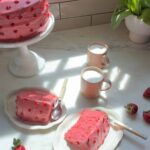 स्ट्रॉबेरी लेयर केक स्लाइस, केक स्टैंड पर केक, और कॉफी के कप।