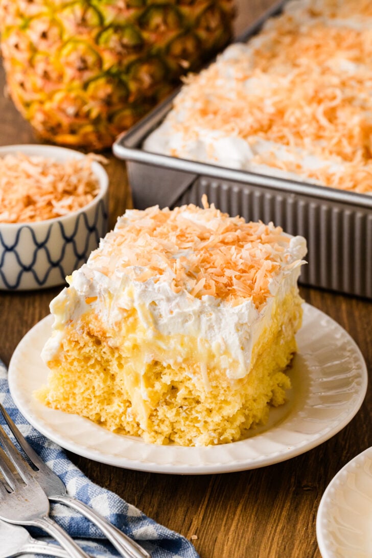 अनानास नारियल पोक केक के ऊपर 13x9 पैन और सफेद प्लेट में व्हीप्ड क्रीम और टोस्टेड नारियल के टुकड़े डालें।